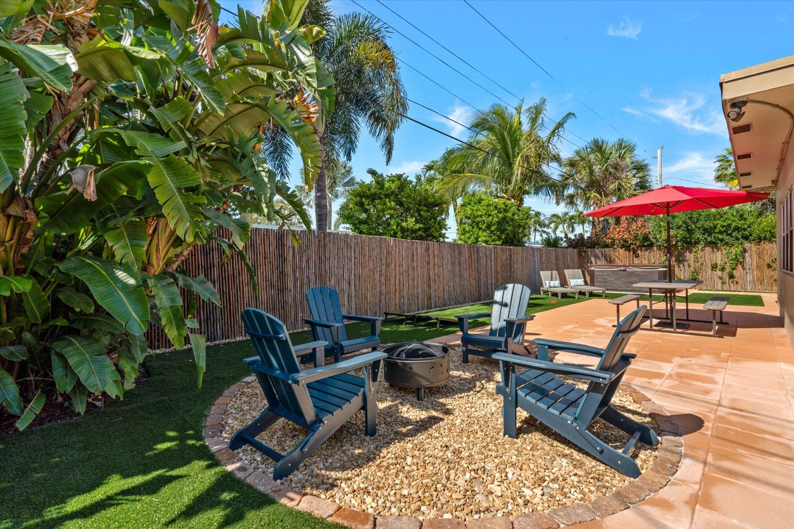 Backyard of 3721 NE 16th Terrace: fire pit circle with Adirondack chairs, paver patio, artificial turf and a bamboo privacy fence under palm trees